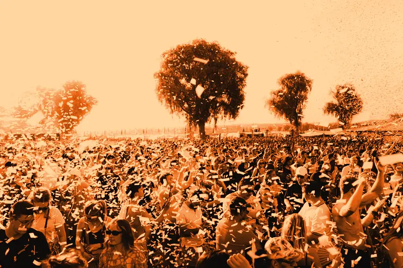 Foule dense qui danse sous une pluie de confettis lors d'un festival en plein air ensoleillé, grands arbres en arrière-plan