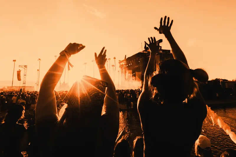 Deux festivalières de dos lèvent les bras au coucher du soleil face à une grande scène en plein air entourée de palmiers