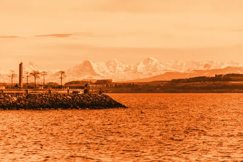 Jetée en enrochement et promenade arborée au bord du lac de Neuchâtel, sommets alpins enneigés à l'horizon