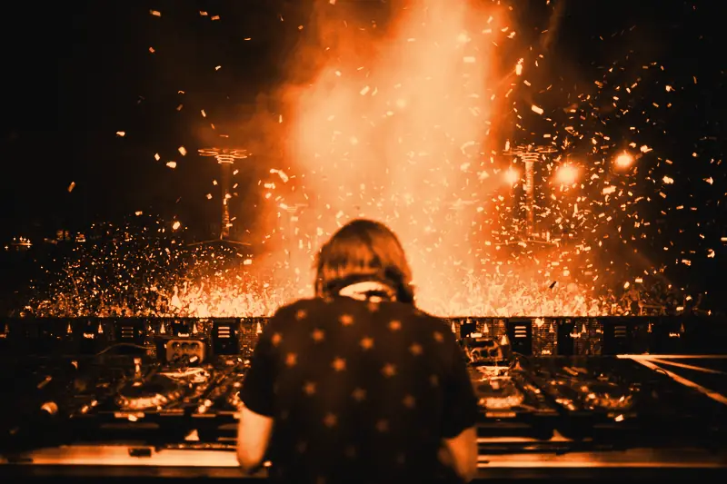 DJ vu de dos derrière ses platines, face à une foule nocturne sous une pluie de confettis et de fumée