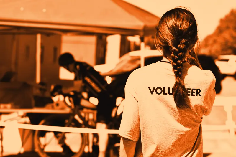 Bénévole aux cheveux tressés, de dos, en t-shirt marqué « Volunteer », près d'une tente d'accueil en plein air